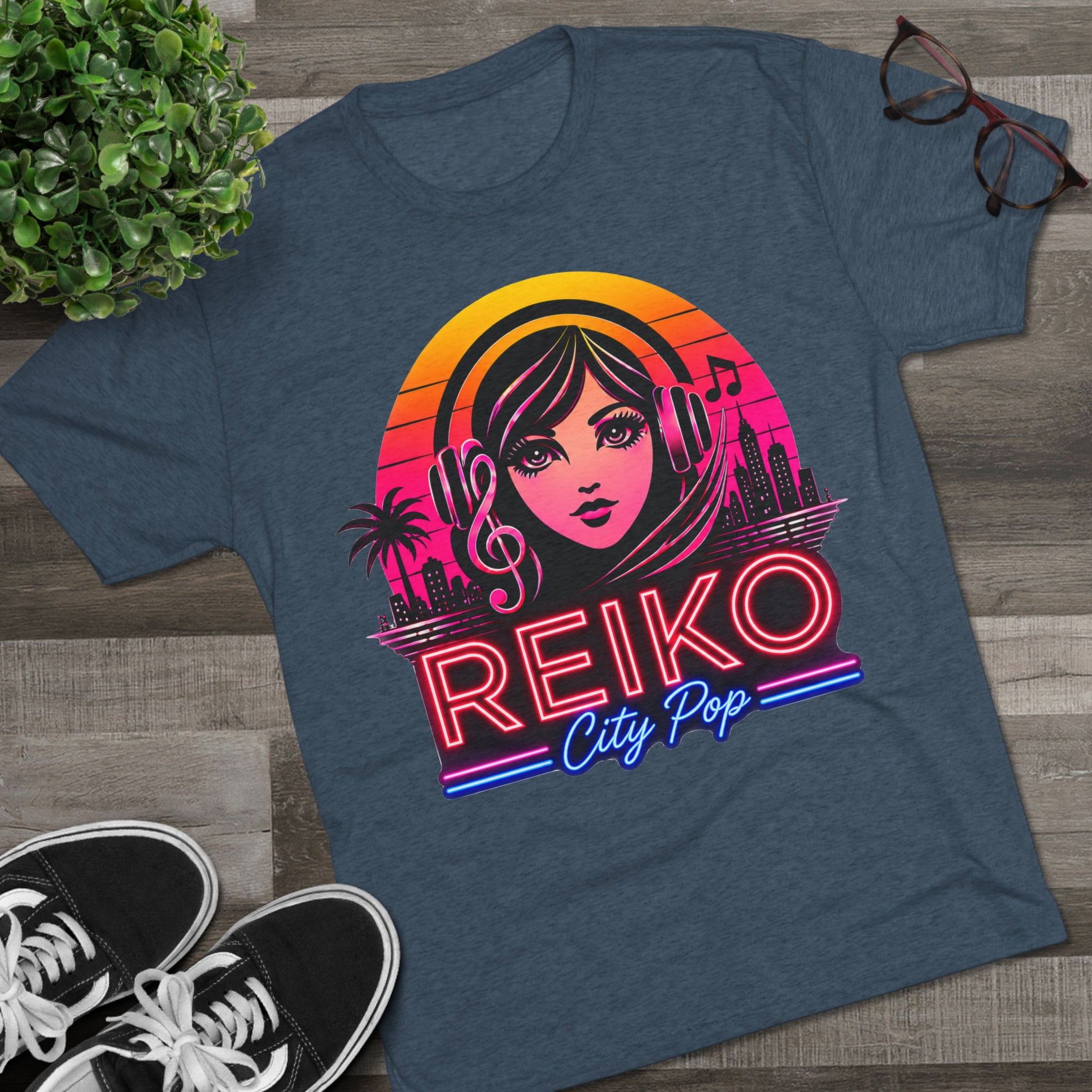 Front view of the Reiko Takahashi City Pop Sunset Logo Tee in Indigo, printed on a premium Next Level tri-blend T-shirt and displayed in a styled flat lay with sneakers, eyeglasses, and a green plant. Features retro neon artwork inspired by the song "Sunset Road."