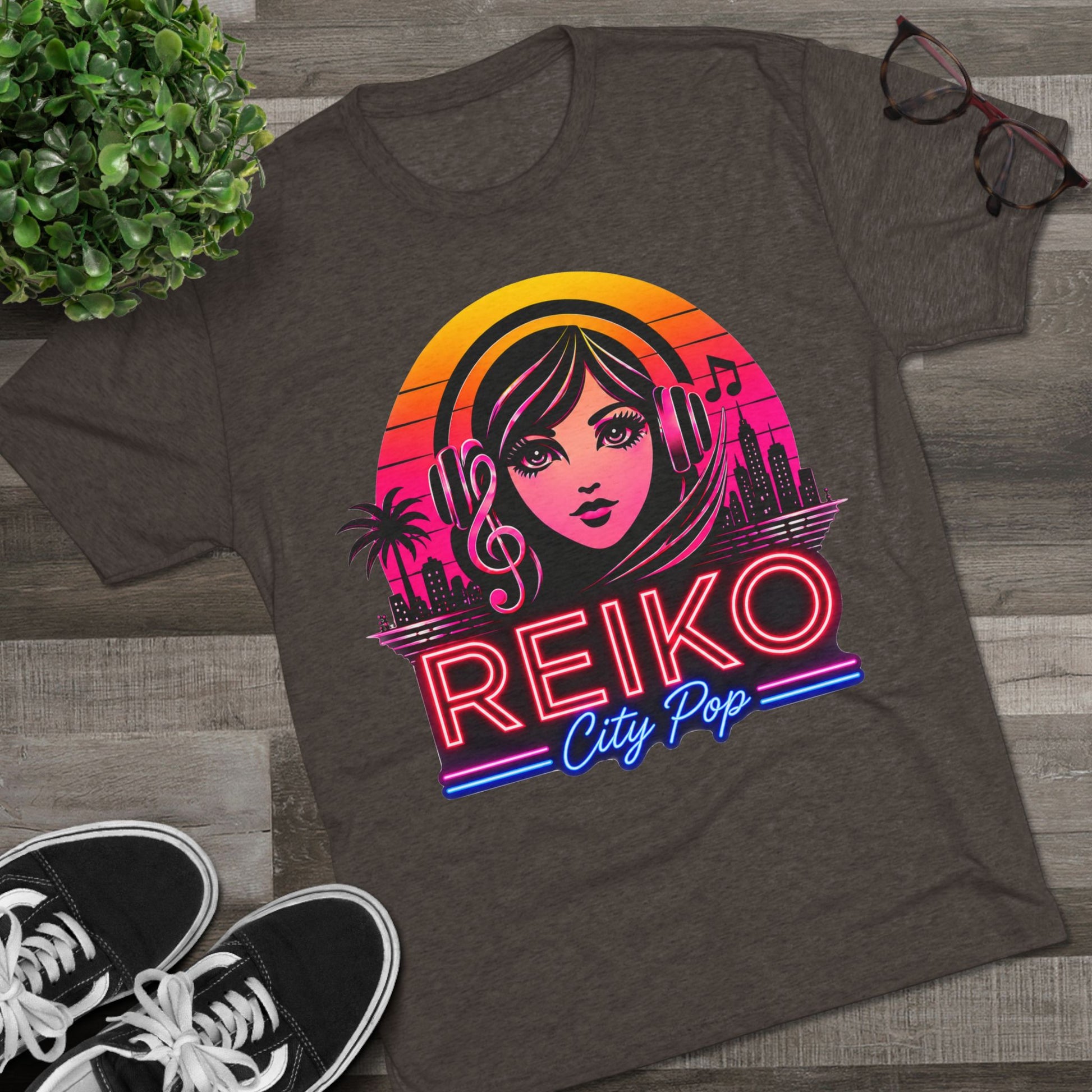 Front view of the Reiko Takahashi City Pop Sunset Logo Tee in Macchiato, printed on a premium Next Level tri-blend T-shirt and displayed in a styled flat lay with sneakers, eyeglasses, and a green plant. Features retro neon artwork inspired by the song "Sunset Road."