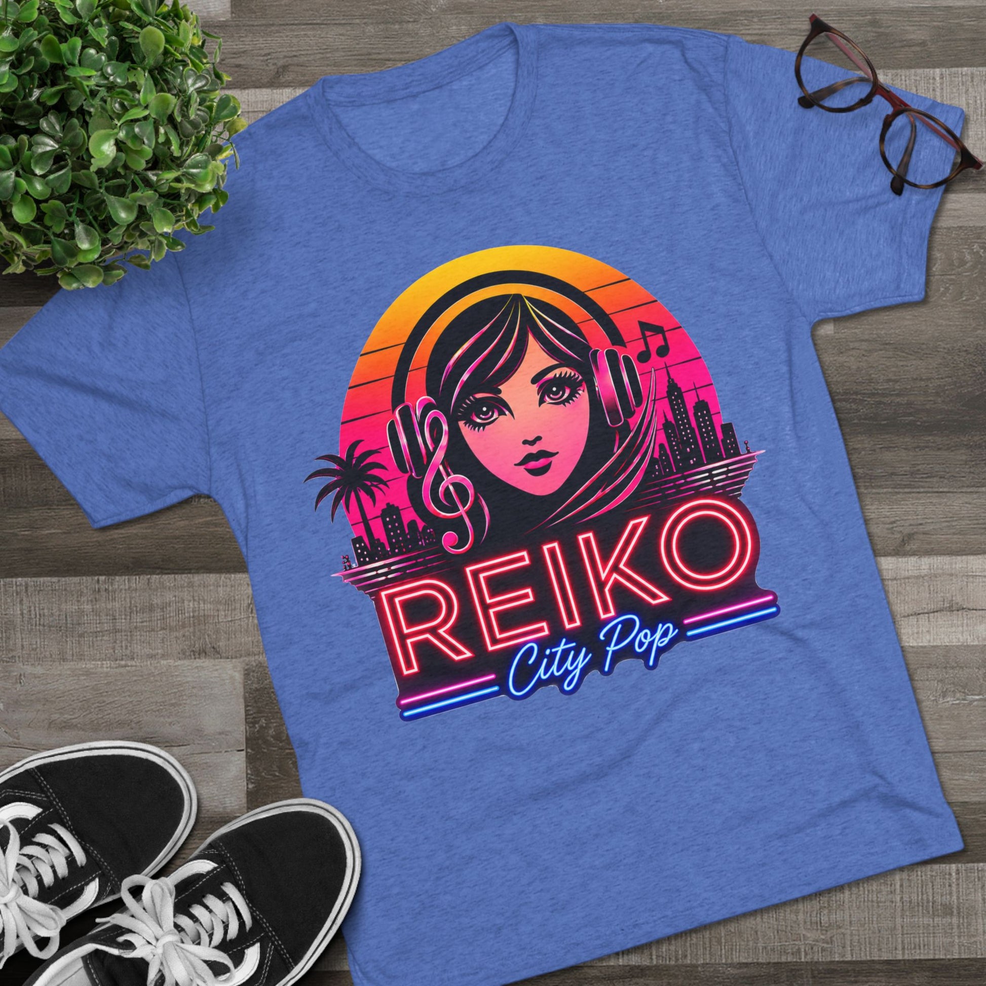 Front view of the Reiko Takahashi City Pop Sunset Logo Tee in Royal Blue, printed on a premium Next Level tri-blend T-shirt and displayed in a styled flat lay with sneakers, eyeglasses, and a green plant. Features retro neon artwork inspired by the song "Sunset Road."